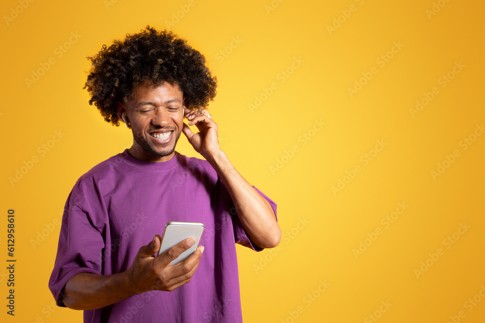 Cheerful black adult curly man in purple t-shirt and wireless headphones looks at phone - obrazy, fototapety, plakaty 
