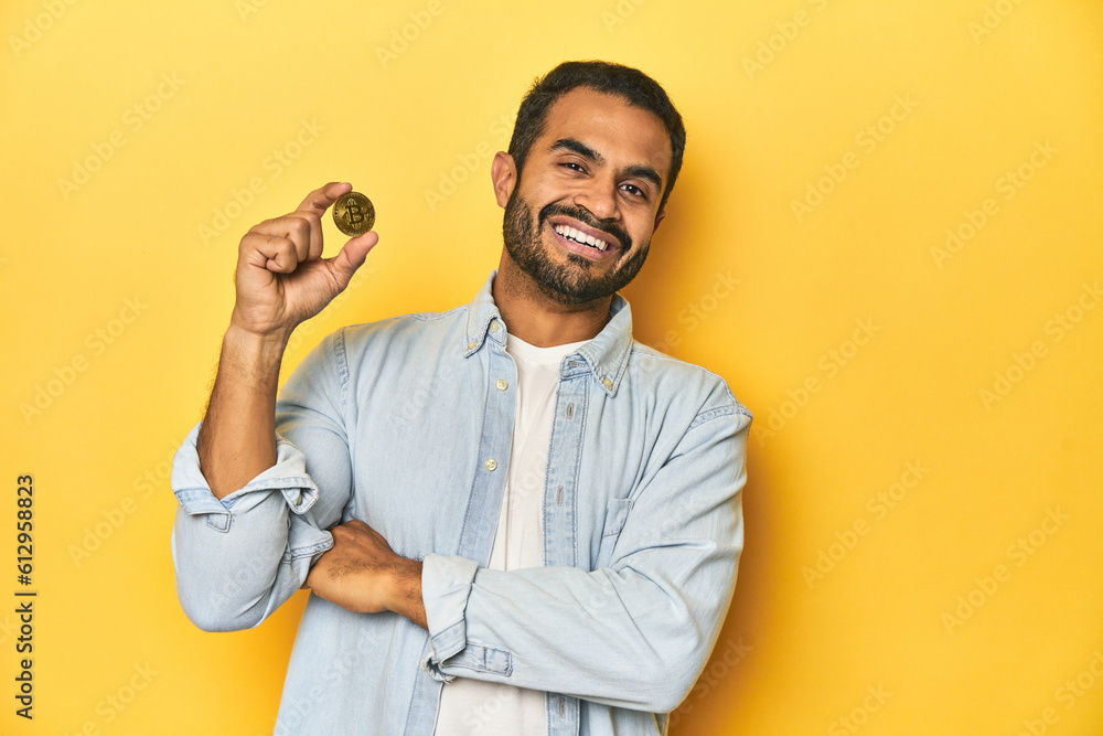 Young Latino man holding a Bitcoin coin, yellow studio background, laughing and having fun. Young Latino man holding a Bitcoin coin, yellow studio background, laughing and having fun.