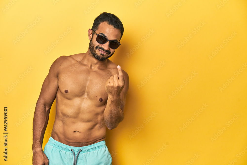 Fit young Latino man in swimwear and sunglasses, yellow studio background, pointing with finger at you as if inviting come closer. Fit young Latino man in swimwear and sunglasses, yellow studio background, pointing with finger at you as if inviting come closer.