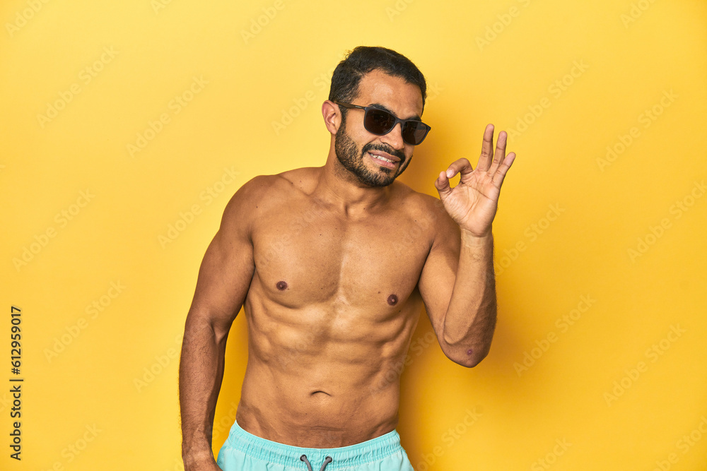 Fit young Latino man in swimwear and sunglasses, yellow studio background, cheerful and confident showing ok gesture.