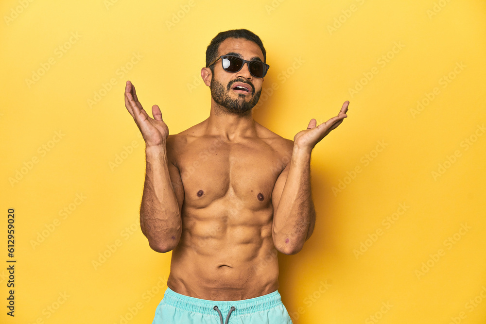 Fit young Latino man in swimwear and sunglasses, yellow studio background, surprised and shocked.