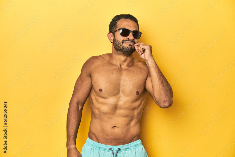 Fit young Latino man in swimwear and sunglasses, yellow studio background, with fingers on lips keeping a secret. Fit young Latino man in swimwear and sunglasses, yellow studio background, with fingers on lips keeping a secret.