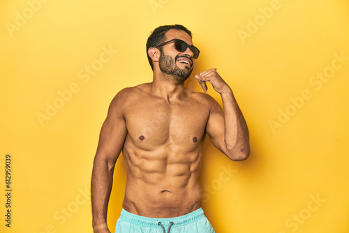 Fit young Latino man in swimwear and sunglasses, yellow studio background, showing a mobile phone call gesture with fingers.