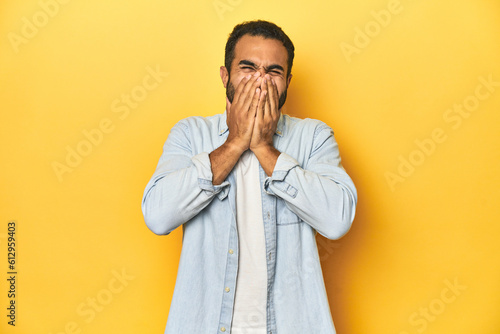 Casual young Latino man against a vibrant yellow studio background, laughing about something, covering mouth with hands.