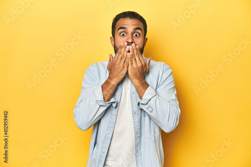 Casual young Latino man against a vibrant yellow studio background, shocked, covering mouth with hands, anxious to discover something new.