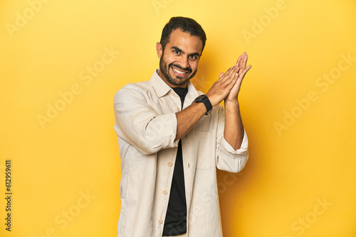 Casual young Latino man against a vibrant yellow studio background, feeling energetic and comfortable, rubbing hands confident.