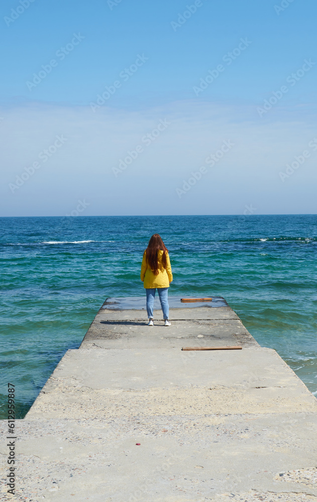 Person young woman in yellow jacket and blue jeans with auburn long hair walking on weat pier, blue sky and the sea.