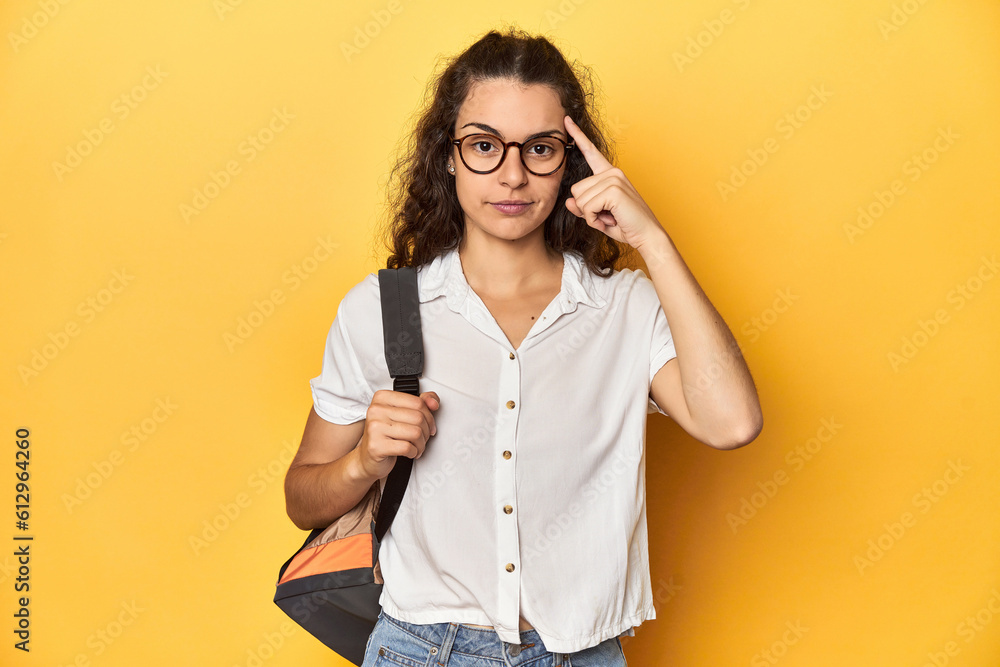 Caucasian university student with glasses, backpack, pointing temple with finger, thinking, focused on a task.