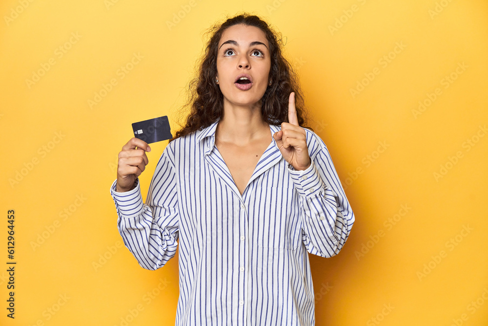 Woman holding a credit card, yellow backdrop, pointing upside with opened mouth. Woman holding a credit card, yellow backdrop, pointing upside with opened mouth.