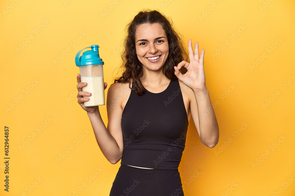 Woman holding a protein shake, in sportswear, cheerful and confident showing ok gesture. Woman holding a protein shake, in sportswear, cheerful and confident showing ok gesture.