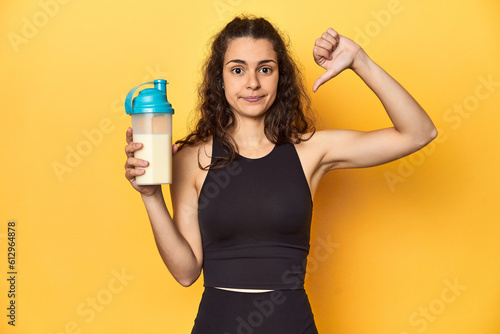 Woman holding a protein shake, in sportswear, showing a dislike gesture, thumbs down. Disagreement concept. © Asier