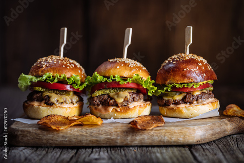 Three burger sliders placed on a wooden board in rustic atmosphere. Very tasty burgers with melted cheese, lettuce, tomato and sauce