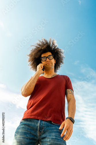 Afro-Styled Man Engrossed in an Energetic Phone Conversation. Charming Afro Guy Expressing with Gusto as he Talks, Backdrop of a Breathtaking Sky. Afro-Textured Man Talking Animatedly on the Phone