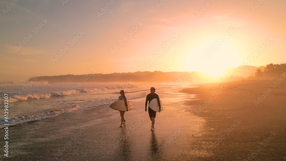 AERIAL: Cheerful surfers admire big waves while walking on sandy shore at sunset