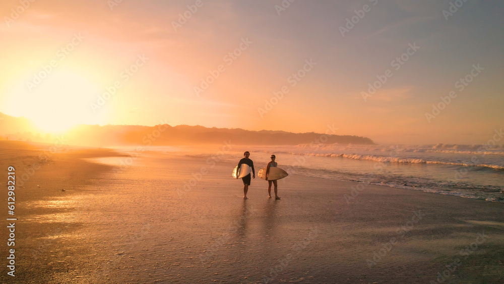 AERIAL: Young couple walking on beautiful sandy beach after sunset surf session