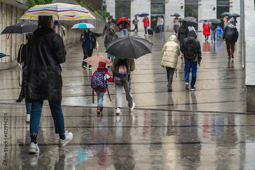 Authentic passers-by in the rain with umbrellas, hoods and no umbrellas walk on a cloudy day along the city street, a crowd of citizens during bad weather, wet passers-by in the city, reflections road photo