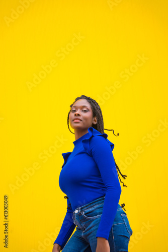 Urban session. Young dark-skinned woman with long braids in a blue dress on a yellow background, with a serious look © unai