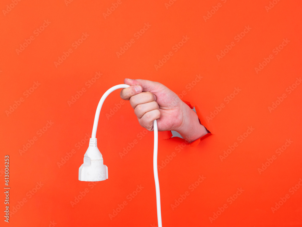 A man's hand holds a with power cable through torn orange paper. Hand through torn orange paper. 
