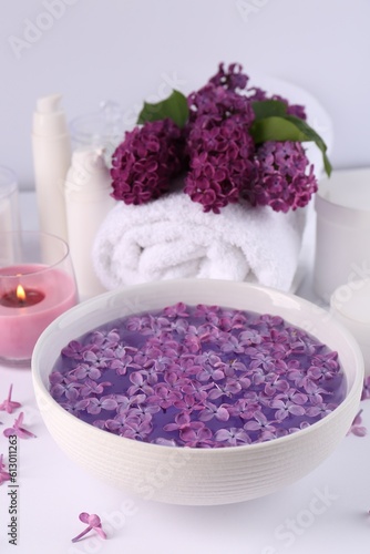 Bowl of water with lilac flowers  spa products and burning candle on white table