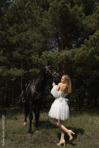 A beautiful caucasian girl in a wedding dress stands next to a horse in the forest