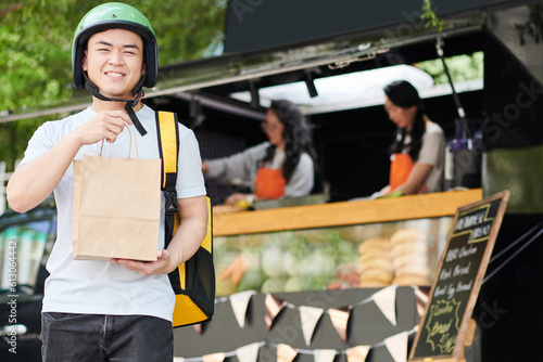 Young smiling courier or deliveryman with paperbag looking at camera while standing against food truck with two women preparing hotdogs