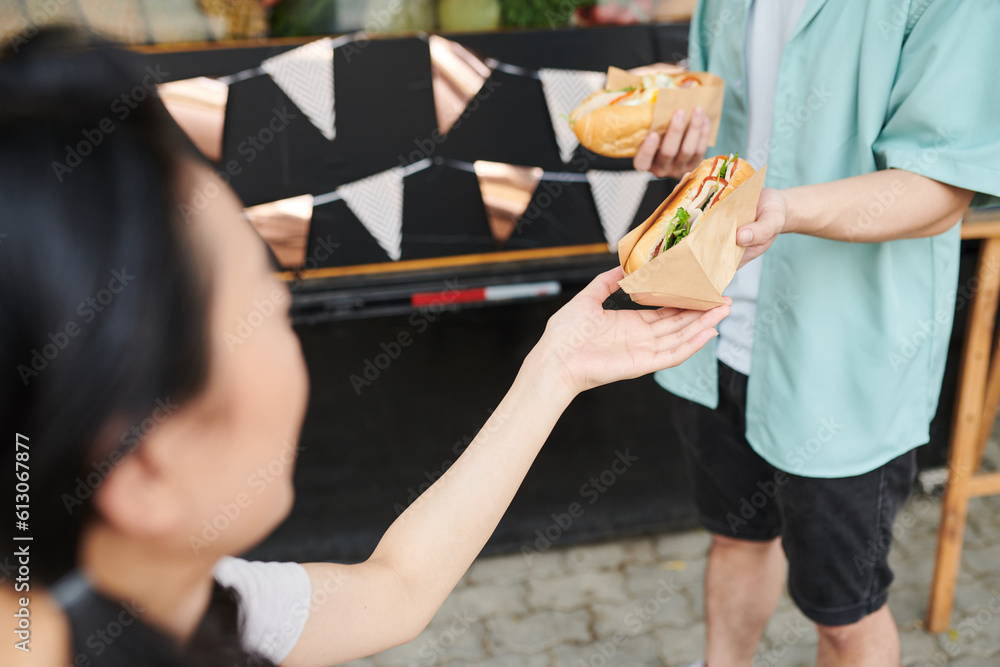 Close-up of young woman taking fresh appetizing hotdog wrapped in paper from hand of her boyfriend in casualwear standing in front