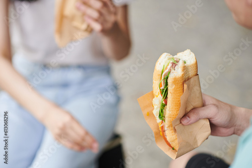 Close-up of fresh appetizing hotdog with vegetables, bacon and cheese held by hungry young man eating it after buying in street food truck