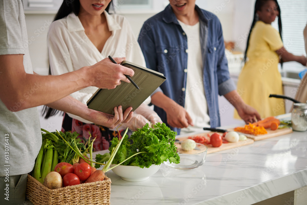 Young man showing online recipe on tablet screen to his wife standing by kitchen table and preparing vegetable salad or other dish with friends Young man showing online recipe on tablet screen to his wife standing by kitchen table and preparing vegetable salad or other dish with friends