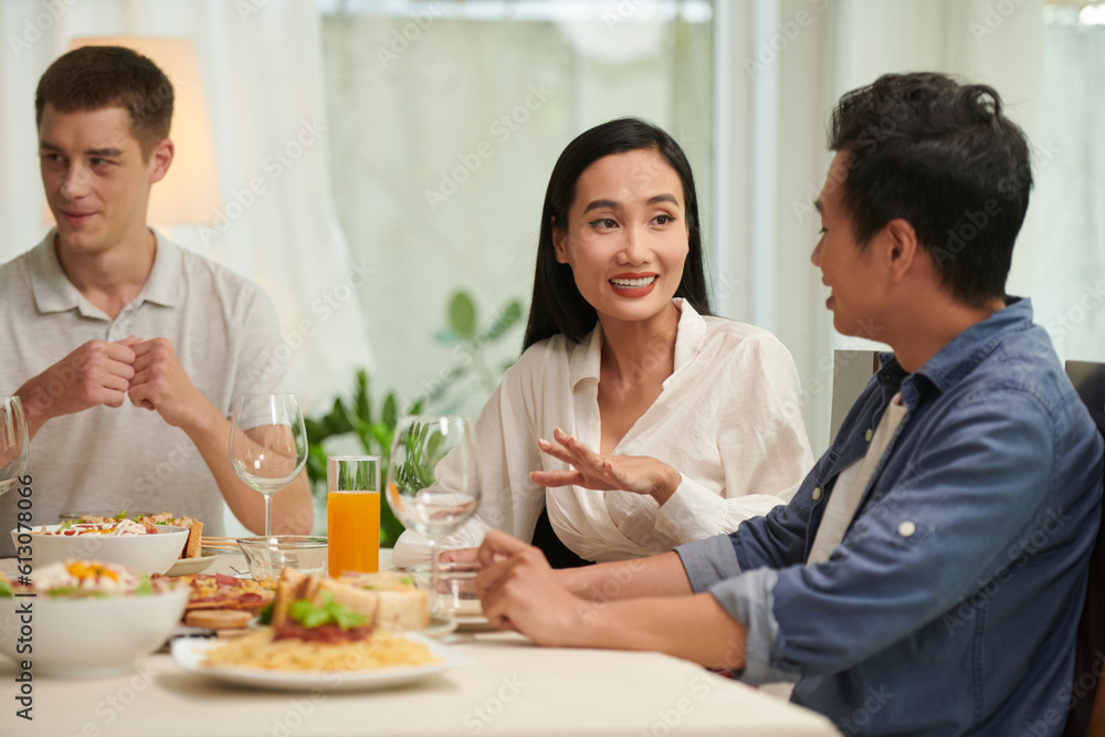 Young smiling Asian woman talking to her boyfriend by table served with appetizing homemade food and drinks for dinner or home party