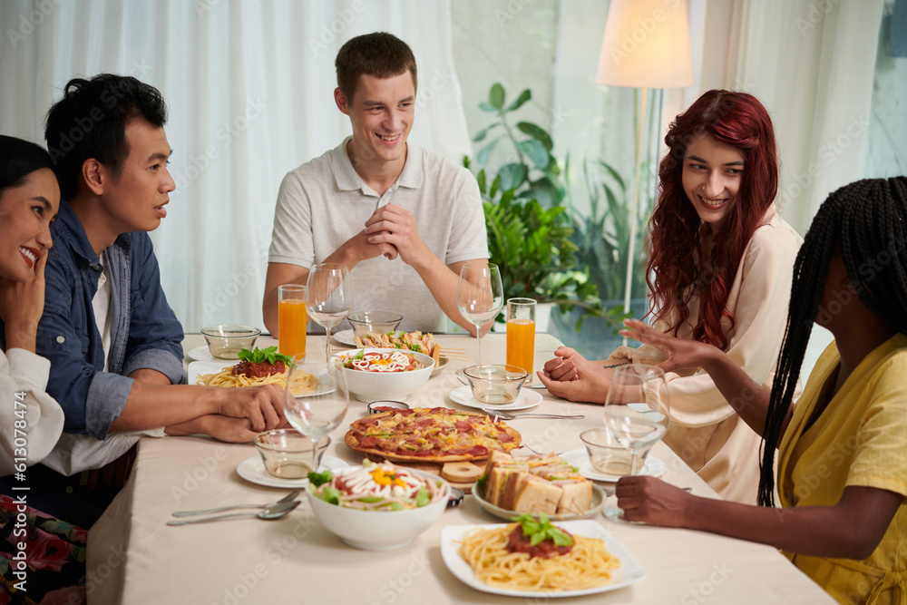 Group of young intercultural people in smart casualwear chatting by served table and enjoying dinner consisting of homemade food Group of young intercultural people in smart casualwear chatting by served table and enjoying dinner consisting of homemade food