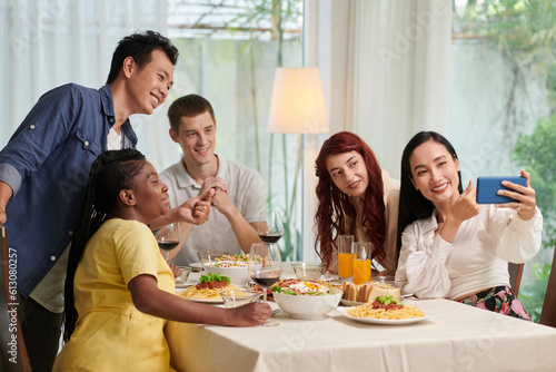 Group of happy young people making selfie or communicating in video chat with their friends while enjoying dinner at home party