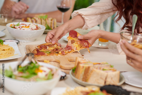 Hands of young hungry woman taking slice of appetizing homemade pizza from wooden board standing on served dinner table with cooked food