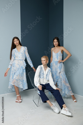 Three fashion models. Asians in identical looks, blue dress with the pattern. Blonde in white sweater, sport pants, trousers with the stripes. Beautiful young women. Bicolor wall, light and dark blue