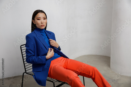 High fashion photo of a beautiful elegant young asian woman in pretty blue jacket, blazer, orange pants, trousers. White textured rounded wall