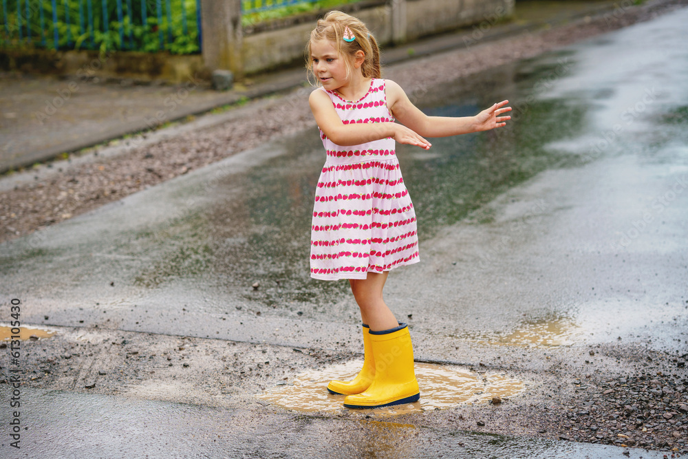 Happy little preschool girl wearing yellow rain boots and walking during puddles. Cute child in colorful clothes jumping into puddle, splashing with water, outdoor activity