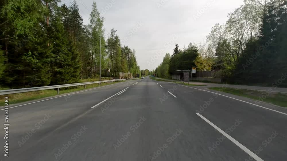 Cars drive along the road with green grass and trees visible on the sides. Houses can also be seen. Cars drive along the road with green grass and trees visible on the sides. Houses can also be seen.