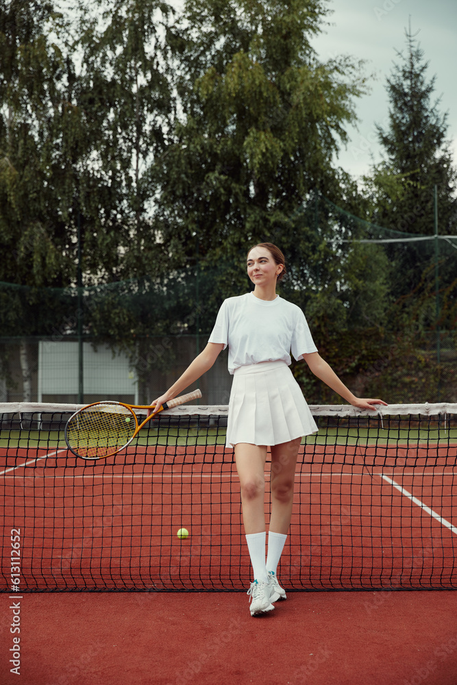 Fashionable female in white clothing with tennis racket posing at tennis net on court. Sports Fashion