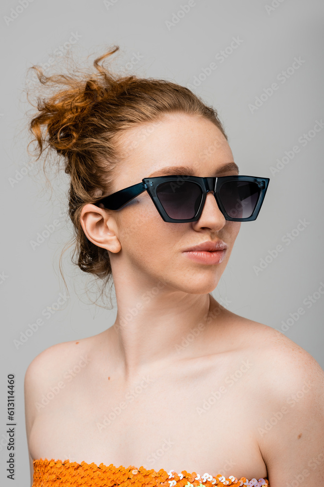 Portrait of young red haired and freckled woman in sunglasses and top with sequins looking away while standing isolated on grey background, trendy sun protection concept, fashion model Portrait of young red haired and freckled woman in sunglasses and top with sequins looking away while standing isolated on grey background, trendy sun protection concept, fashion model