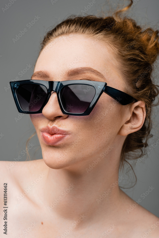 Close up view of young redhead and freckled woman in stylish sunglasses with naked shoulders pouting lips and standing isolated on grey background, trendy sun protection concept, fashion model Close up view of young redhead and freckled woman in stylish sunglasses with naked shoulders pouting lips and standing isolated on grey background, trendy sun protection concept, fashion model