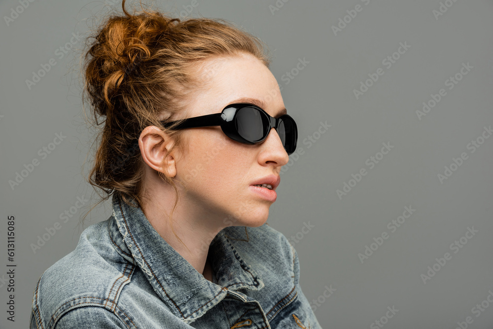 Fashionable young red haired woman with natural makeup wearing sunglasses and denim jacket while standing isolated on grey background, trendy sun protection concept, fashion model Fashionable young red haired woman with natural makeup wearing sunglasses and denim jacket while standing isolated on grey background, trendy sun protection concept, fashion model