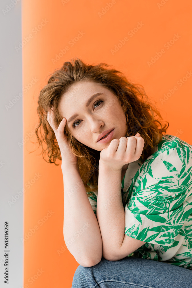 Portrait of young redhead woman in blouse with floral pattern and jeans looking at camera while standing on orange and grey background, trendy casual summer outfit concept, Youth Culture Portrait of young redhead woman in blouse with floral pattern and jeans looking at camera while standing on orange and grey background, trendy casual summer outfit concept, Youth Culture