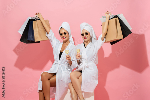 Two woman friends wearing spa bathrobes holding shopping bags and drinks in pink color isolated background studio shot