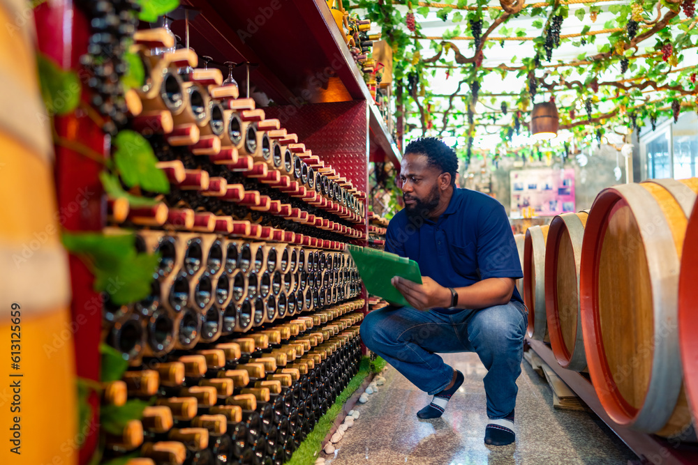 Naklejka premium African man liquor shop worker working and counting a bottle of wine, champagne or beer on shelf in store warehouse. Winery manufacturing industry, winemaker and alcoholic drink business concept.