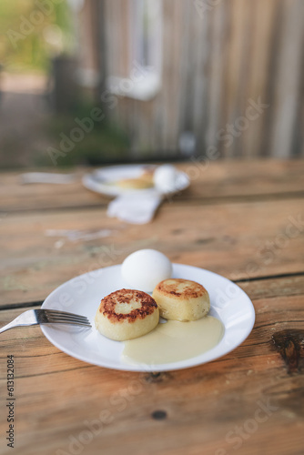 Two white plates with cheesecakes, sweet sauce, eggs and metal fork on aged wooden table. Village atmosphere and early morning breakfast for two persons, with old village house background.