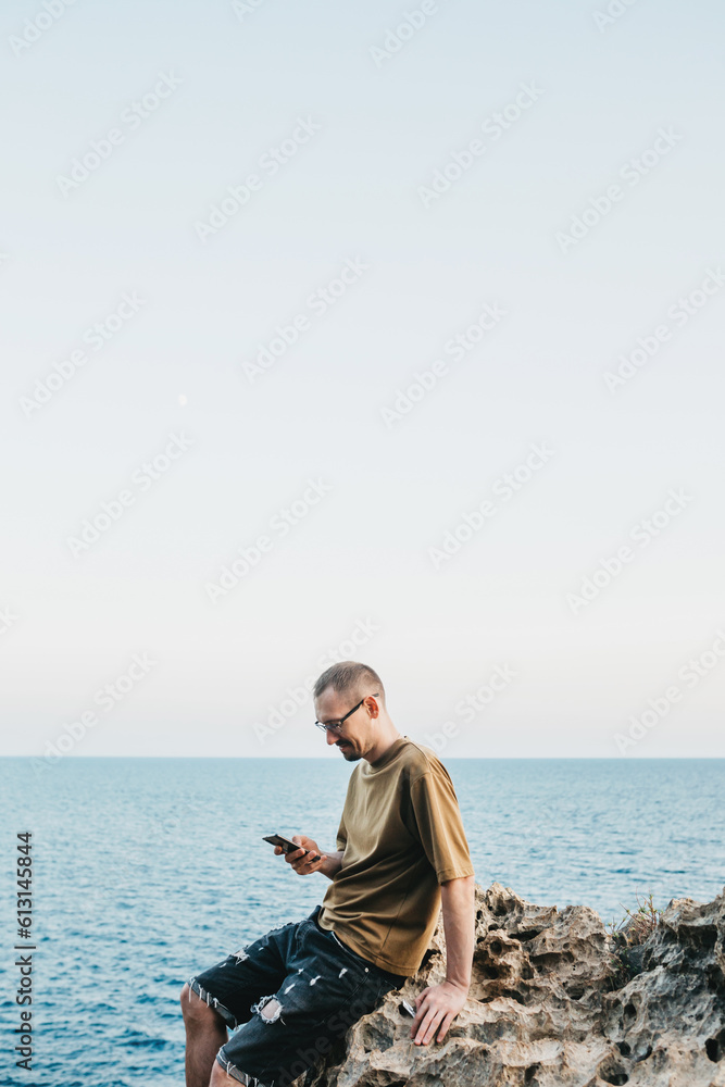 A happy young man is sitting alone on the seashore. A man enjoys the beautiful nature of the sea around him and writes a message on his phone A happy young man is sitting alone on the seashore. A man enjoys the beautiful nature of the sea around him and writes a message on his phone