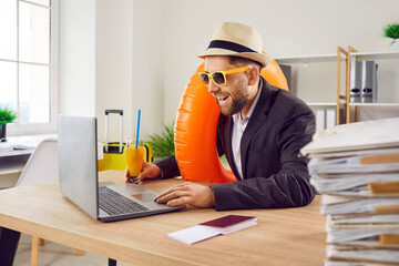 Funny happy business man sitting at the desk of his workplace in office with juice cocktail, boarding pass and passport holding rubber ring and booking tickets for summer vacation online via laptop.