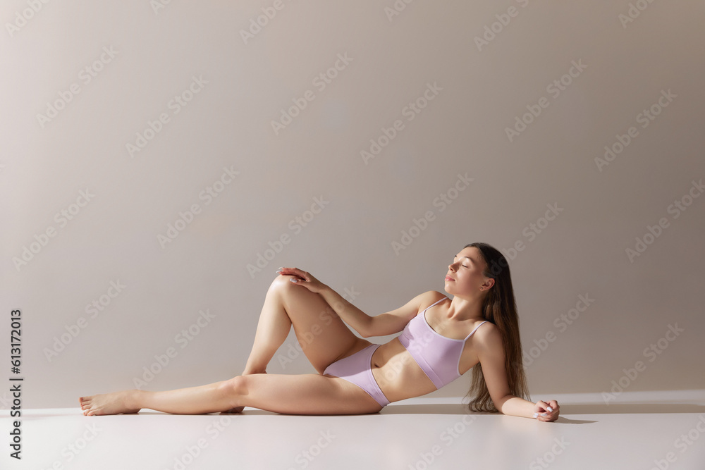 Fototapeta premium Relaxed young girl with fit, slim figure lying on floor, posing in underwear against grey studio background