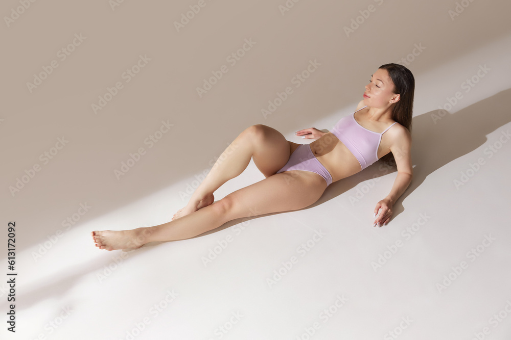 Top view young girl with fit, slim figure lying on floor, posing in underwear against grey studio background. Tenderness