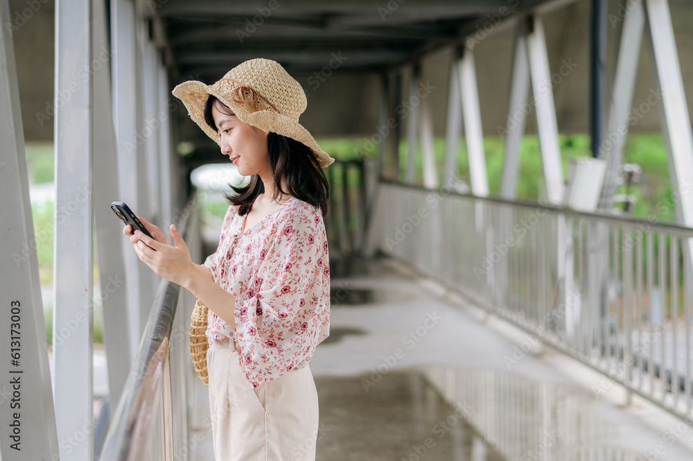 young asian woman traveler with weaving basket using mobile phone and standing on overpass. Journey trip lifestyle, world travel explorer or Asia summer tourism concept.