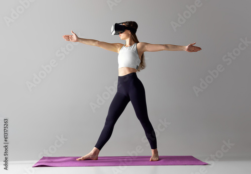Young girl with slim body, wearing VR glasses and training in sportswear against grey studio background. Online sport exercises